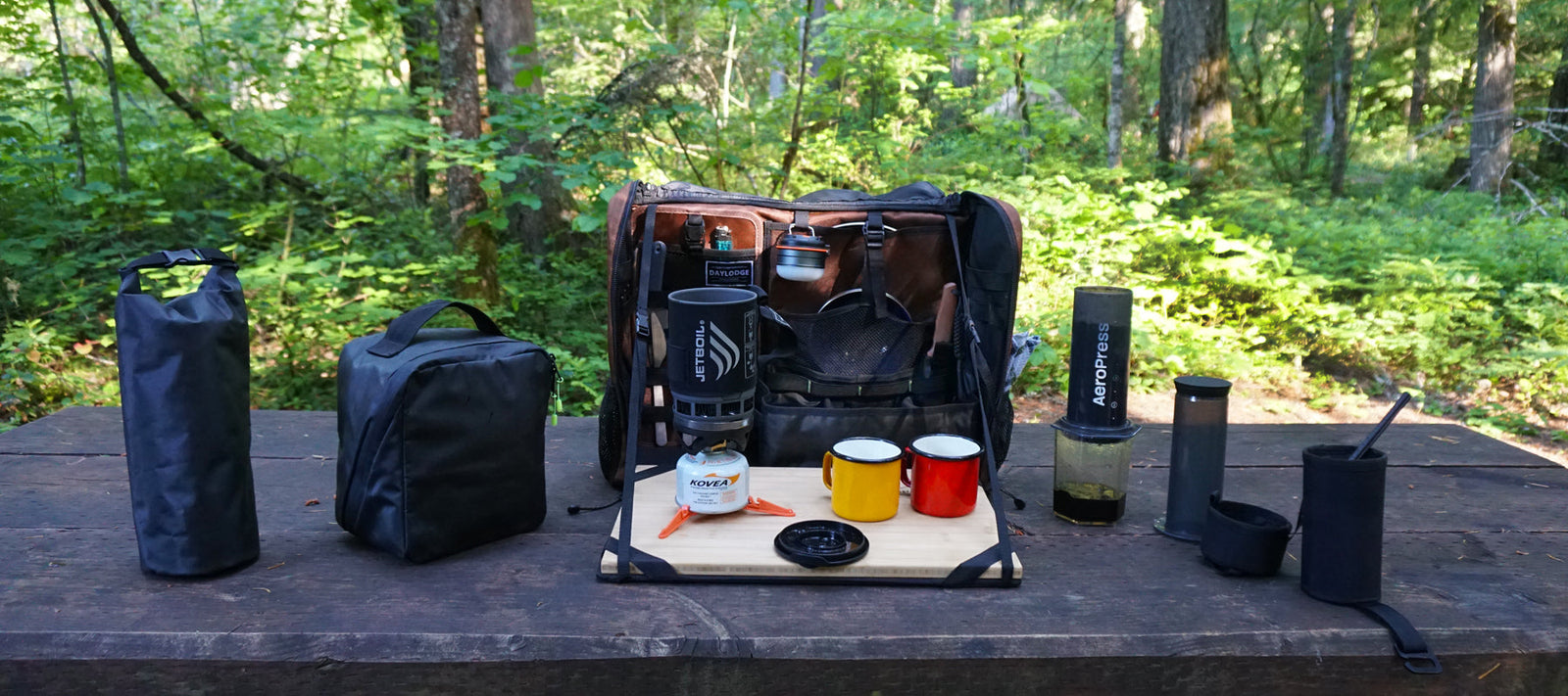 The Workspace bag open on a table in the woods with a French press and two orange mugs on the bamboo surface