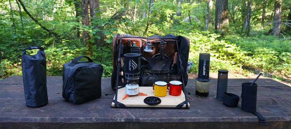 The Workspace bag open on a table in the woods with a French press and two orange mugs on the bamboo surface