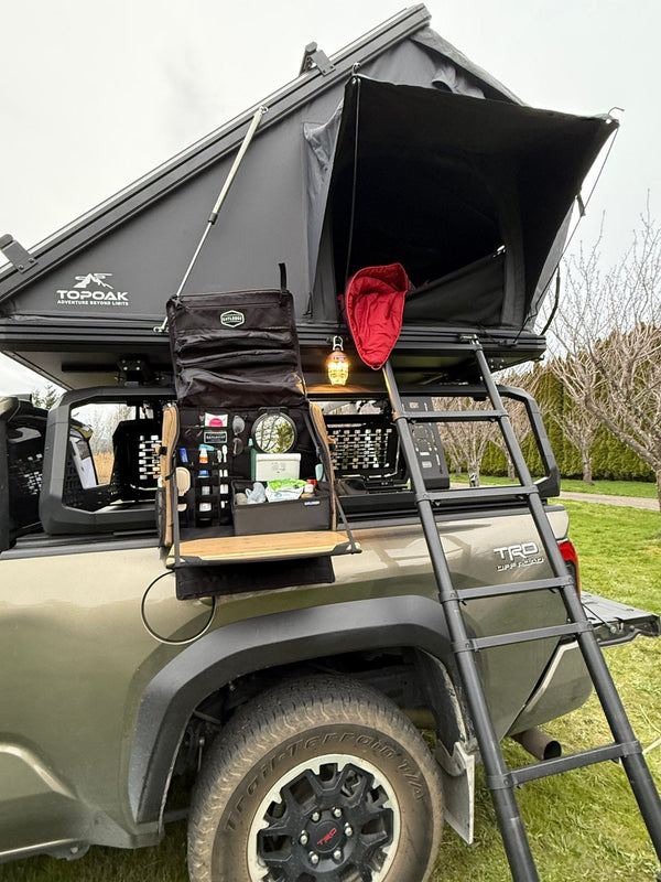 Workspace bag hung open on a truck bed rack beside the rooftop tent ladder, set up as a camp vanity