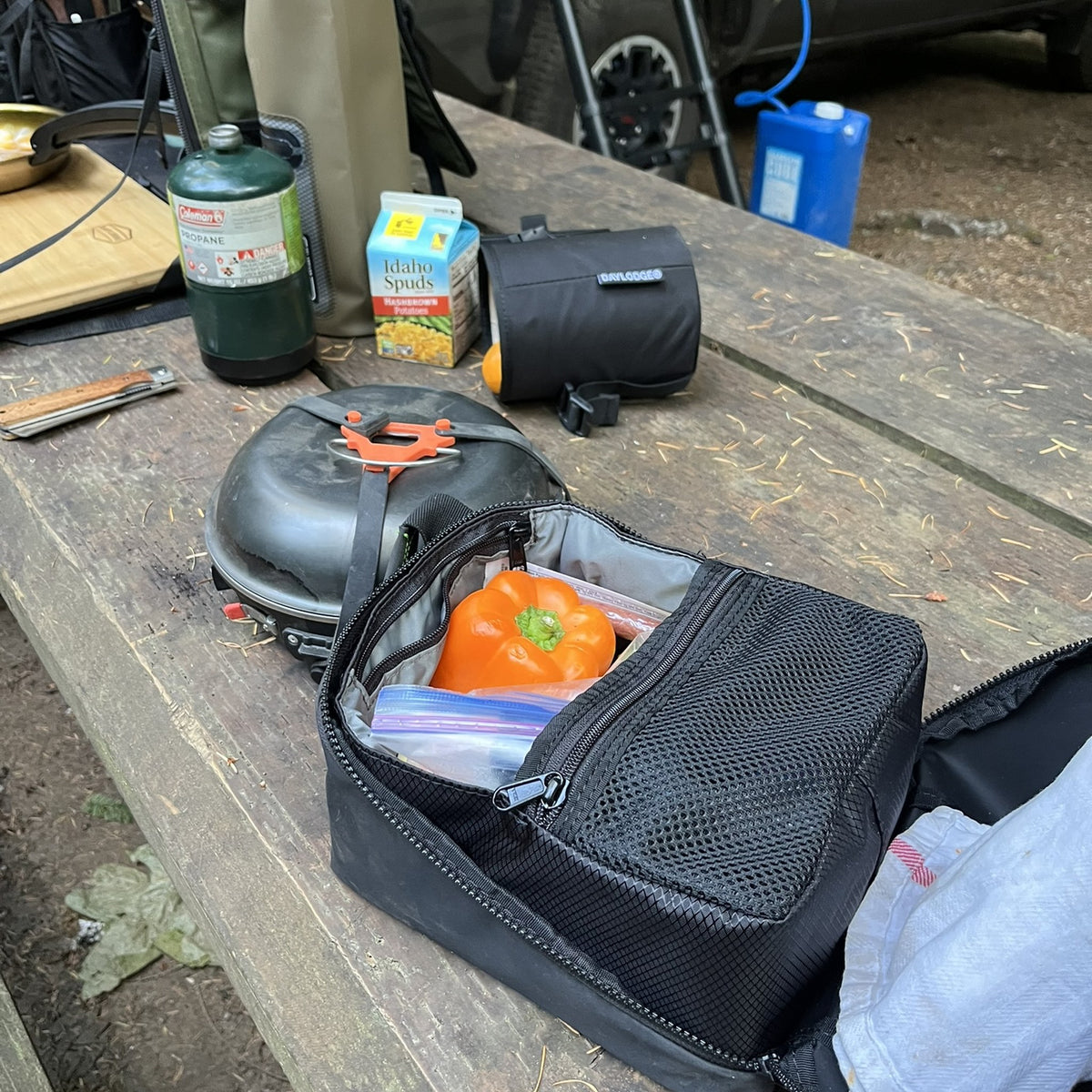 Cold Cube soft cooler open on a campsite picnic table, packed with produce, next to a Workspace bag