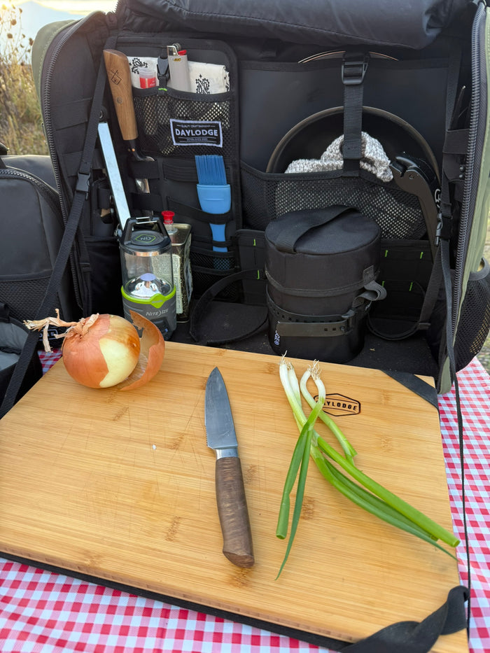 Workspace open on a gingham picnic table with an onion, knife and green onions on the bamboo board